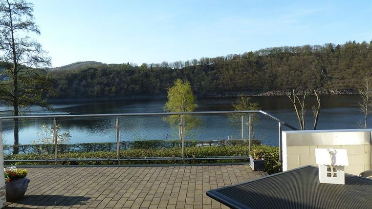 A tranquil lake with green shores and gentle hills in the background. On the terrace in the foreground, there are plants and a table.