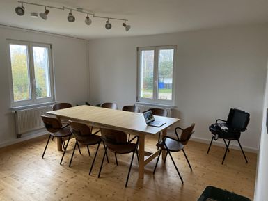 Ein heller Meetingraum mit einem großen Holztisch und mehreren Stühlen. Auf dem Tisch steht ein Laptop und es gibt große Fenster, die viel Tageslicht hereinlassen.