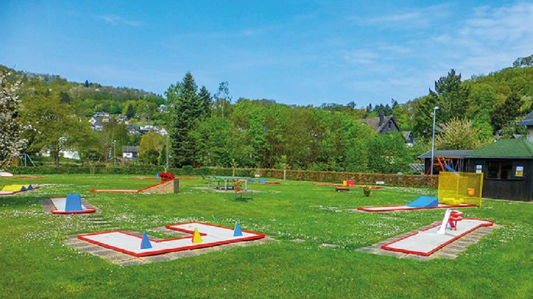 Een minigolfbaan in een groen park, omgeven door bomen en heuvels. De spelers kunnen op verschillende holes spelen.