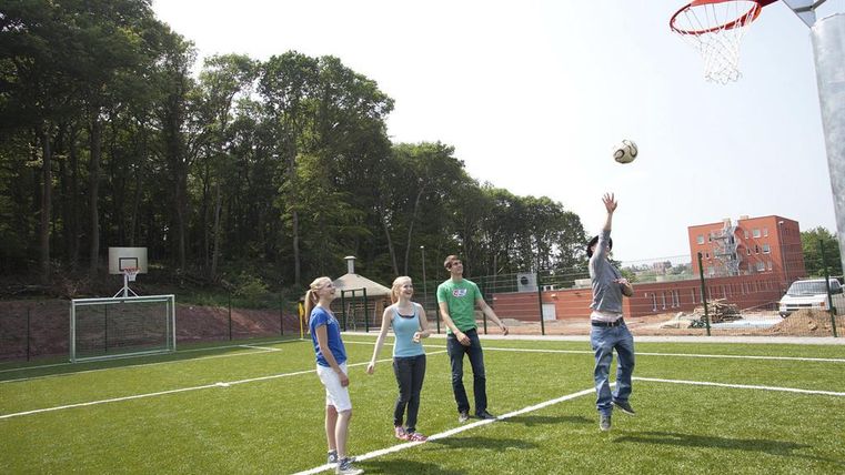 A group of young people is playing basketball on a sports field with artificial turf. In the background, basketball hoops and trees are visible.