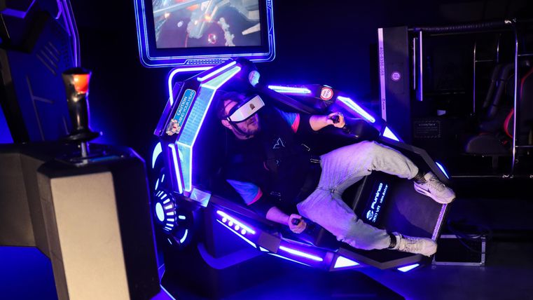 A person is sitting in a modern VR simulator with glowing lights. Interactive elements are visible on the screen, showcasing an immersive experience.