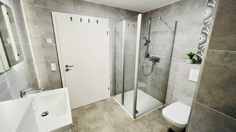A modern bathroom with gray tiles. It features a shower, a sink, and a toilet.