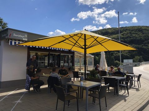 Een gezellig café buiten met een grote gele parasol. Gasten zitten aan tafels en genieten van de zon.