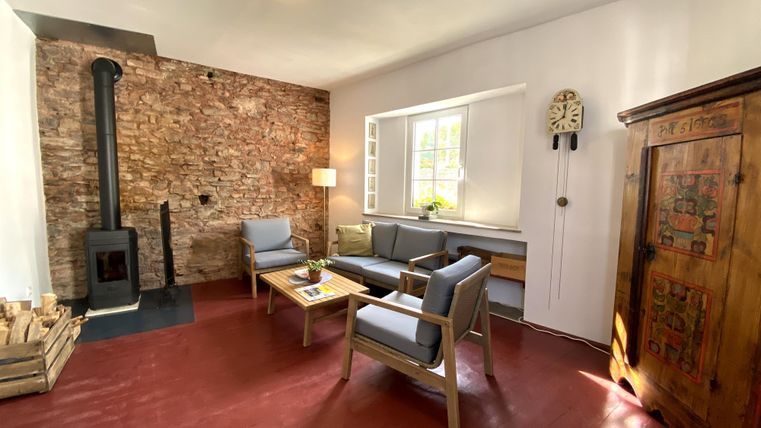 Een gezellige woonkamer met een houten open haard, blauwe fauteuils en een rustieke kast. De muren zijn gedeeltelijk met stenen bekleed en er komt veel natuurlijk licht binnen.