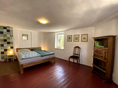 Ein gemütliches Schlafzimmer mit einem Doppelbett und einem Holzstuhl. Die Wände sind hell, und es gibt kunstvolle Bilder und sanfte Beleuchtung.