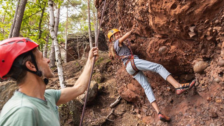 A climber hangs on a rock face while a person safeguards him. Both are wearing helmets and are surrounded by trees.