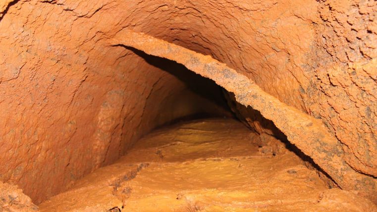Ein enger Tunnel mit rötlich-braunen Wänden und einer rauen Oberfläche. Der Boden ist aus einem mineralischen Material, das die Wände eingeklemmt erscheint.