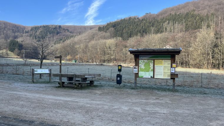 A quiet place with an information stand and a bench in nature. In the background, gentle hills and a blue sky can be seen.