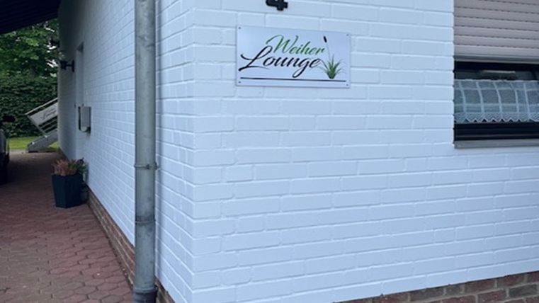 Een wit huis met een lichte wand en een klein bord met de tekst "Weberei Lounge". Naast de ingang staat een groen plantenbak.