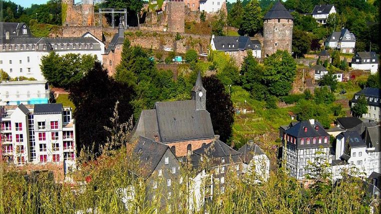 Eine malerische Stadtlandschaft mit historischen Türmen und gepflegten Gebäuden. Umgeben von grünen Bäumen und sanften Hügeln.