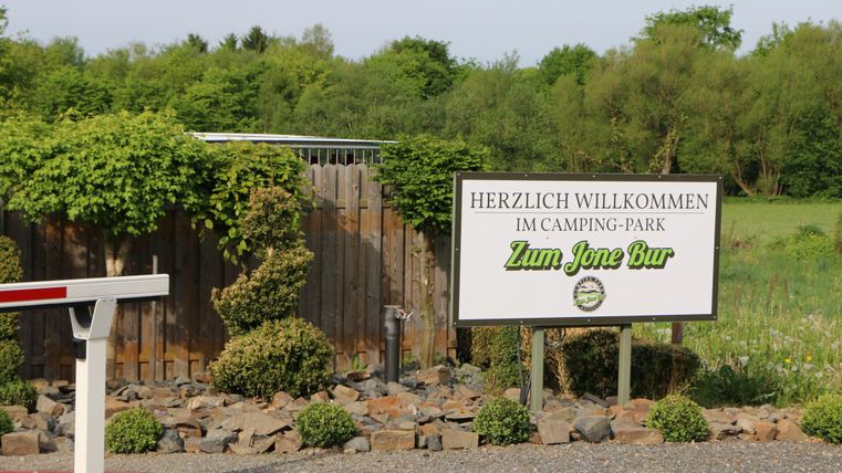 A welcome sign in front of the camping park "Zum Jonen Bur". The surroundings are green and covered with trees and shrubs.
