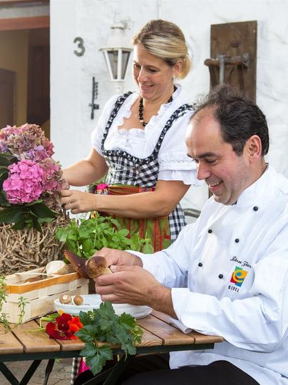 A cook is preparing fresh ingredients while a woman in a traditional costume dress is arranging flowers. In the background, herbs and vegetables are spread out on a table.