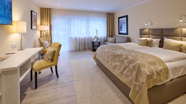 Ein elegantes Hotelzimmer mit einem großen Doppelbett und zeitloser Einrichtung. Viel Licht fällt durch die Fenster, und ein Schreibtisch mit Stuhl ergänzt den Raum.