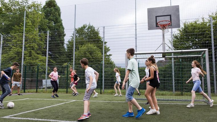 A group of children is playing soccer on a sports field. In the background, a basketball hoop can be seen.