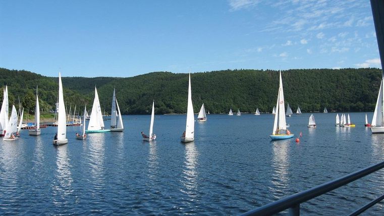 A peaceful lake landscape with many sailboats. Green hills in the background and a blue sky complement the scenery.