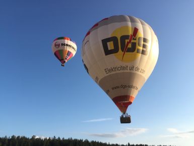 Zwei Heißluftballons schweben über einer Waldlandschaft. Der größere Ballon trägt das Logo einer Firma und strahlt in der Abendsonne.