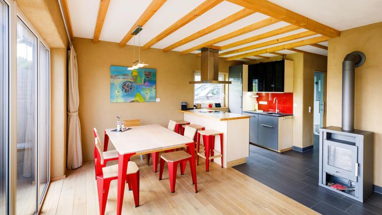 A modern kitchen with a large dining area and red chairs. The room design is bright and friendly, with wooden beams on the ceiling.