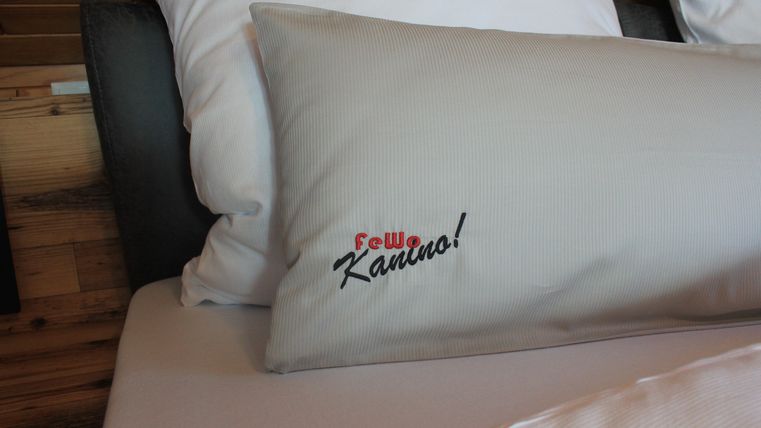 A pillow with the print "Frau Kanuro!" lies on a bed. The background features a natural wood look.