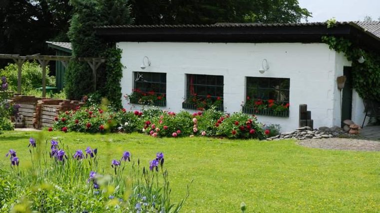 Een charmant, wit huis met kleurrijke bloemen bij het raam. De tuin is weelderig groen, met veel bloeiende planten.