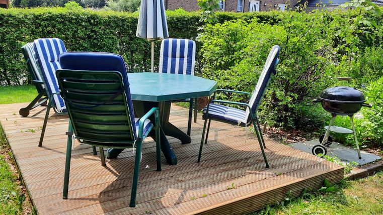 An inviting garden area with a round table and chairs on a wooden terrace. In the background, there is a grill and lush greenery.