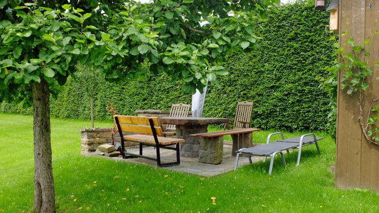 An inviting garden with a stone table set and several chairs. The surroundings are enclosed by a tall hedge and there is a lounger for relaxing.
