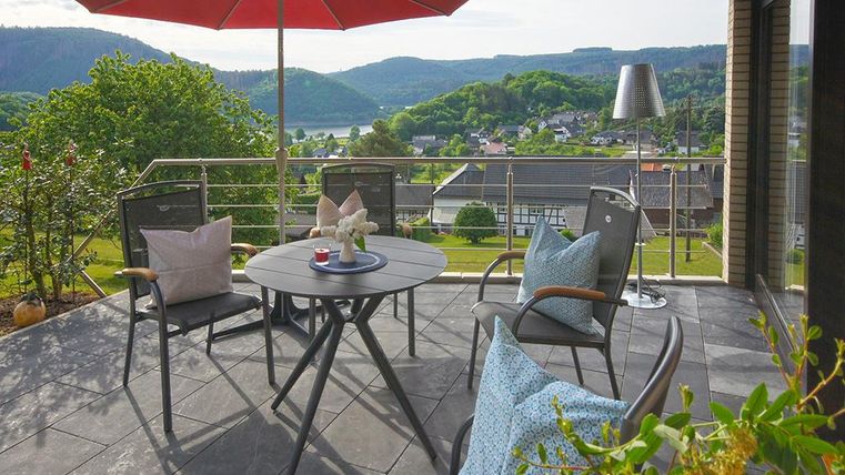 Eine gemütliche Terrasse mit Blick auf grüne Hügel. Ein runder Tisch und Stühle laden zum Entspannen ein, während ein roter Sonnenschirm Schatten spendet.
