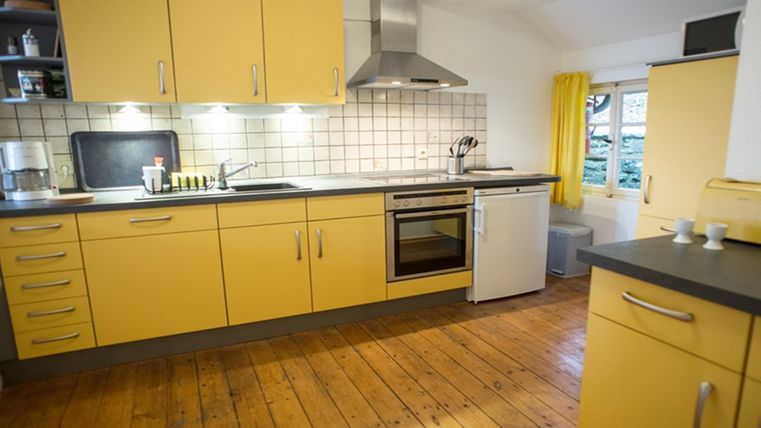 A modern kitchen with yellow cabinets and wooden flooring. It is well-equipped and has large windows with curtains.