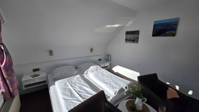A cozy bedroom with two beds and a small table. The walls are decorated with pictures and there is plenty of natural light.