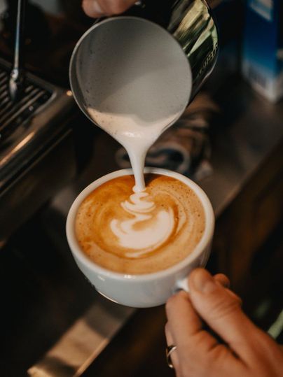 A person pours steaming milk foam into a cup of coffee. The artistic latte art is clearly visible.