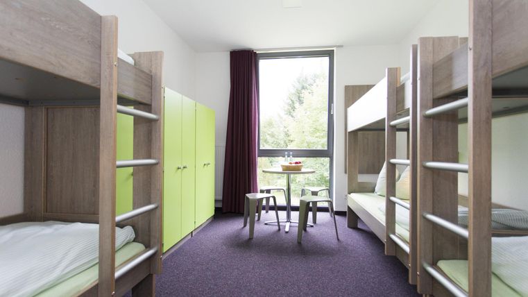 A modern room with two bunk beds and a small table. Large windows let in a lot of light and there are green cabinets.