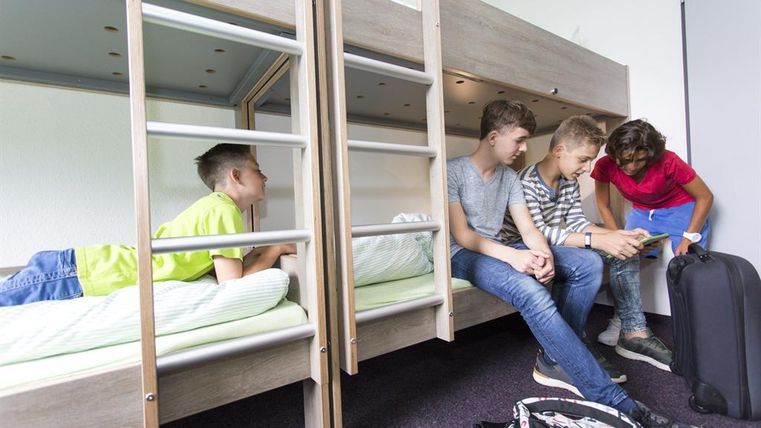 A group of four teenagers in a dormitory. Two are sitting on a lower bed, while another is lying on the bed and showing something to a person.