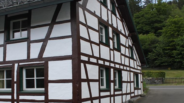 A traditional half-timbered house with white walls and dark beams. In the background are trees and a peaceful environment.