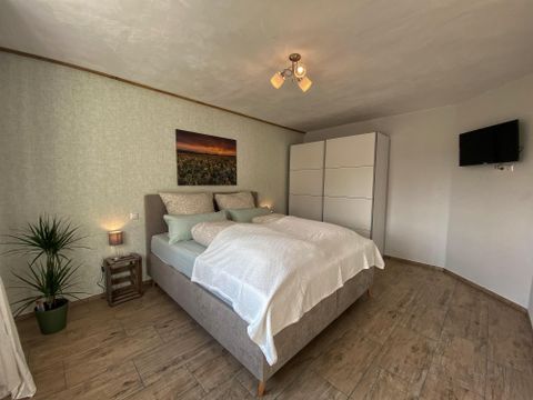 A cozy bedroom with a large bed and bright pillows. A picture hangs on the wall and there is a television and a plant in the room.