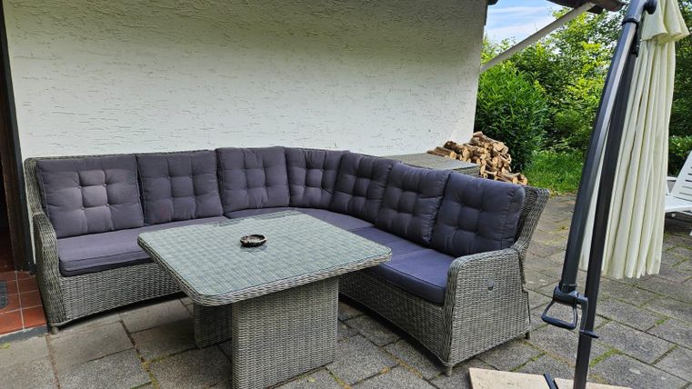 Eine moderne Außensitzgruppe aus Rattan mit grauen Kissen und einem quadratischen Tisch. Der Bereich ist in einem ruhigen Garten mit einem gemütlichen Ambiente.