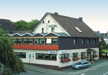 Een hotelgebouw met een beschutte buitenruimte en een blauwe lucht. Voor het hotel staat een blauwe wagen en er zijn bomen in de omgeving.
