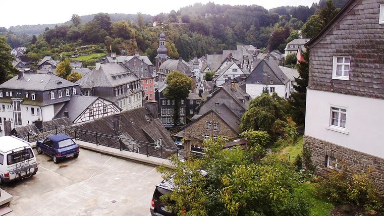 A picturesque view of the town with traditional houses and dense trees. In the background, hills and a church can be seen.