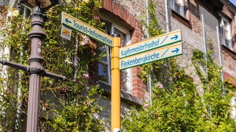 Ein Wegweiser zeigt die Richtung zu verschiedenen Orten, einschließlich der Tourist-Information und der Finkenbergkirche. Umgeben von einer grünen Pflanze und Backsteinmauern vermittelt die Szene ein einladendes Gefühl.