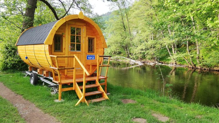 Ein gemütliches Holzhäuschen steht am Ufer eines ruhigen Gewässers, umgeben von grünen Bäumen. Der Weg neben dem Haus führt zum Wasser und bietet einen schönen Ausblick.