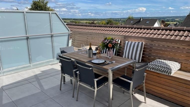 Een gezellige terrasse met een eettafel en stoelen. Op de tafel staan bloemen en een fles wijn, met uitzicht op het landschap.