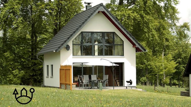 A modern holiday home surrounded by green meadows and tall trees. It features a bright facade with large windows and an inviting terrace.