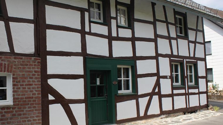 A traditional half-timbered house with white walls and brown beams. The door is green and the floor is paved with stones.