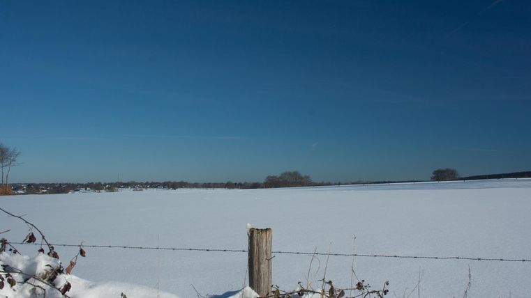 A snow-covered landscape under a clear blue sky. In the foreground, there is a wooden post with a fence.