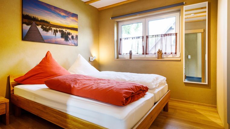 A cozy bedroom with a double bed and colorful pillows. A beautiful landscape picture hangs on the wall, and there is a window with plenty of natural light.