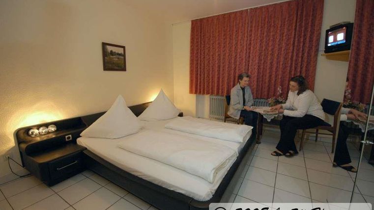 Een eenvoudige hotelkamer met een tweepersoonsbed en twee personen die aan de tafel zitten. De muren zijn versierd met rode gordijnen en er is een televisie in de kamer.