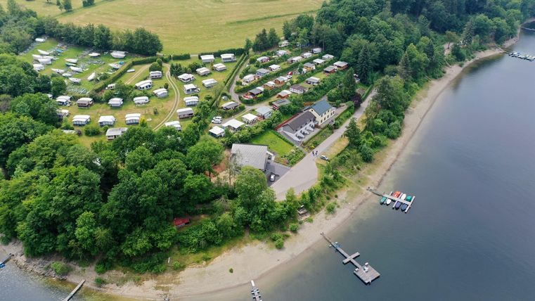 A beautiful campsite by the shore of a body of water, surrounded by trees. Several tents are scattered across the grounds, and there are docks for boats.