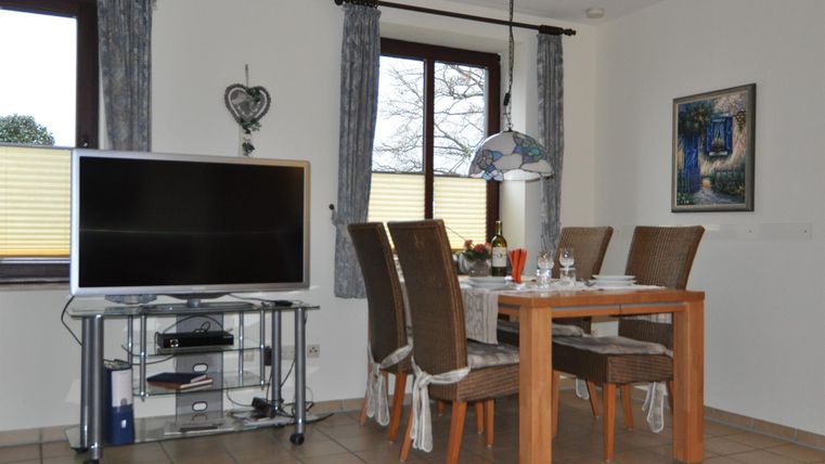 A modern dining area with a wooden table and upholstered chairs. A large television stands on a table, and natural light falls through the windows.
