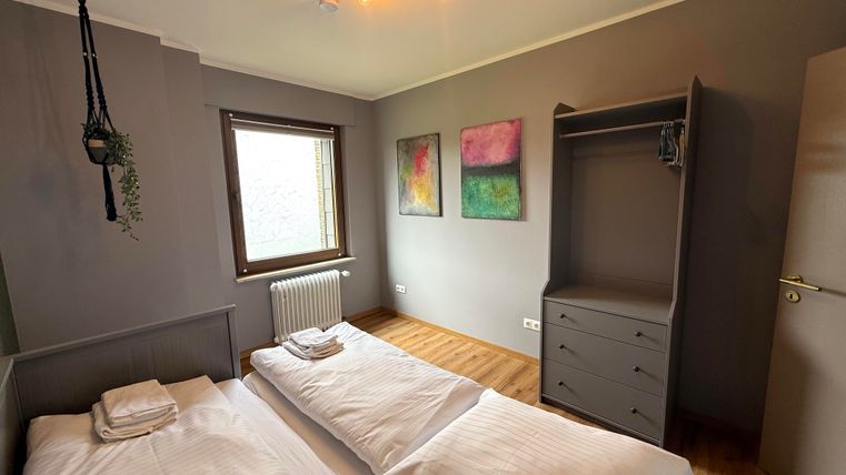 Een gezellige slaapkamer met twee eenpersoonsbedden en een houten vloer. De muren zijn grijs, gedecoreerd met moderne schilderijen, en er is een kast en een raam voor daglicht.