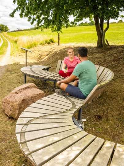Een moderne, gebogen zitgelegenheid in de openlucht. Twee personen zitten samen en praten in een groene landschap.