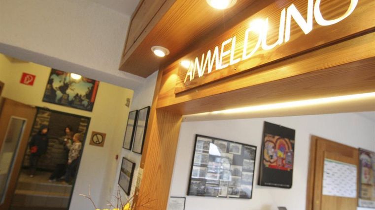 Een ontvangstruimte met een houten balie en de tekst "AANMELDING". Op de achtergrond zijn gasten te zien en enkele wanddecoraties.