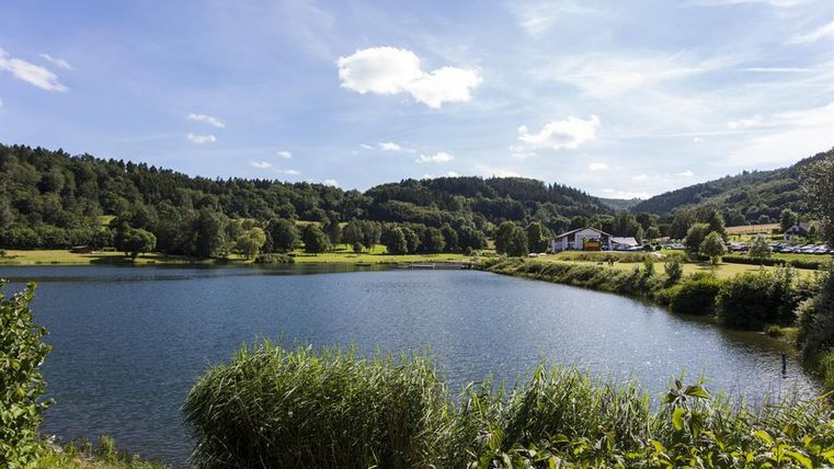 A tranquil lake surrounded by green meadows and gentle hills. The sky is blue and adorned with a few white clouds.